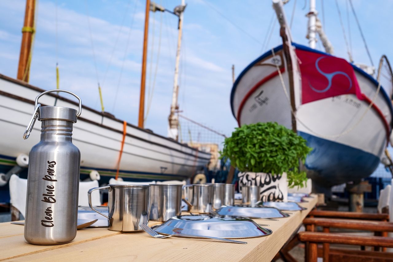 Photo down a long table of reusable bottles and campaign materials, with a sailing boat in the background