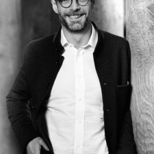 Profile photo for Adonis Lemos, Executive advisor at Common Seas. He wears a dark blazer and white shirts, plus glasses. He smiles at the screen. Photo is in greyscale.