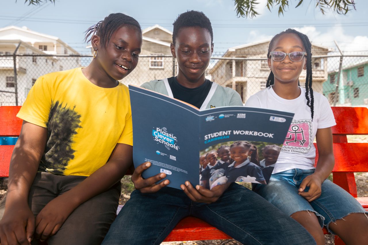 Three Bajan children sit reading the Plastic Clever Schools workbook.