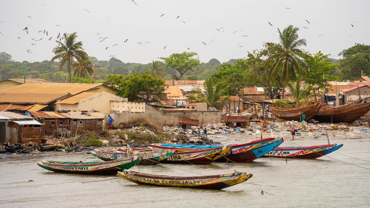 An aerial photo of the Gambian coast looking towards boats and palm trees. The beach is covered with brightly-coloured plastic pollution.