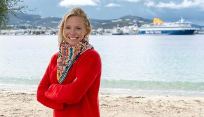 A woman - Jo Royle - in a red sweater standing on a beach with the sea in the background