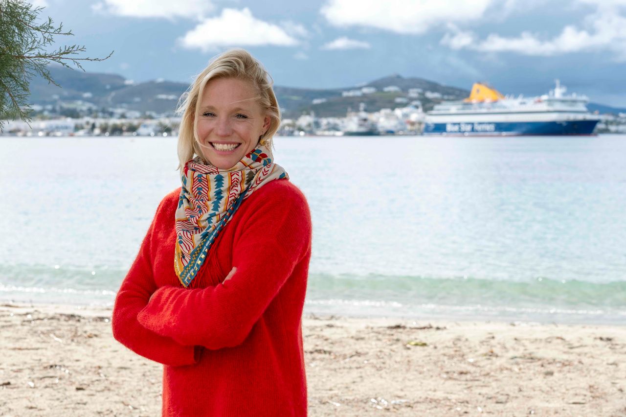 A woman - Jo Royle - in a red sweater standing on a beach with the sea in the background