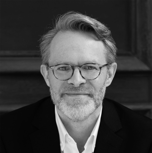 Profile photo of James Simpson, Head of Communications and Marketing at Common Seas. James wears glasses, a white shirt and dark blazer and smiles at the camera. Photo is in greyscale.