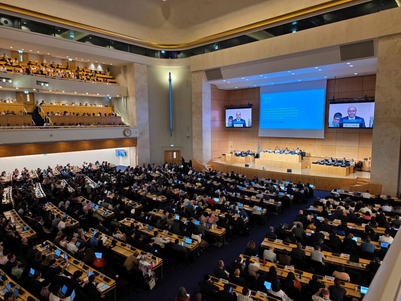 INC5.2 Global Plastics Treaty closing plenary session. The Assembly Hall in the UN's Palais des Nations shot from a high angle, with delegates seated and a screen showing the session.