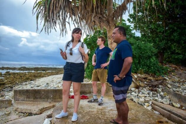 Common Seas team and local partner by coast in Tuvalu. Three people stand on a stone platform under a palm tree with the sea and rocky beach in the background. Plastic waste is visible on the ground.