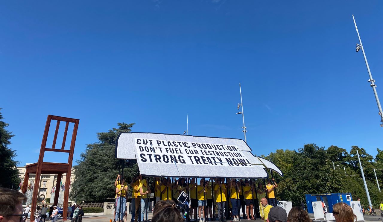 Civil society mobilisation plastics treaty - group of demonstrators at the Place des Nations in Geneva. Demonstrators hold placards that positioned together form a giant speech bubble reading 'cut plastic production, don't fuel our destruction, strong treaty now/'. The Broken Chair is visible in the background against a deep blue sky.