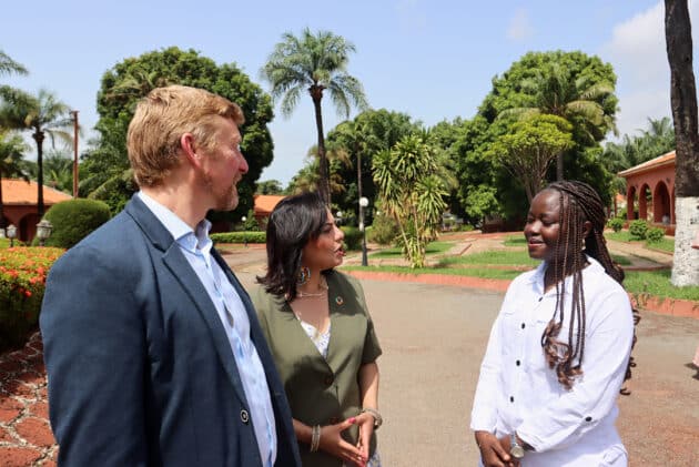 Three people talking outside in Bissau, Guinea-Bissau. Palm trees and other greenery in background by a road.